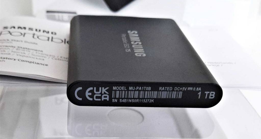 Eine schwarze, getestete externe SSD-Festplatte von Samsung, mit Fokus auf die Modellnummer liegt neben der Gebrauchsanleitung auf der weißen Verpackung.
