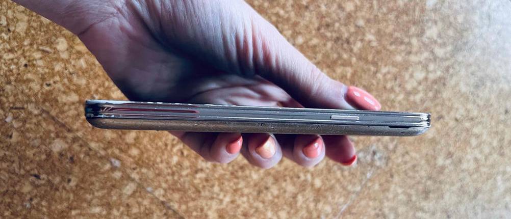 Hand hält ein kleines Smartphone im Test, mit Fokus auf die schmale lange Seite inklusive Einschalttaste, vor braun gesprenkeltem Hintergrund.