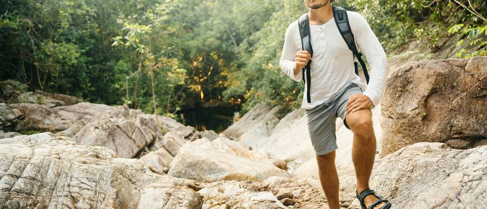Sandalen im Test: Ein Mann wandert über kleine Steinhügel und hat Wanderhut, Shirt, Shorts und Sandalen an. 