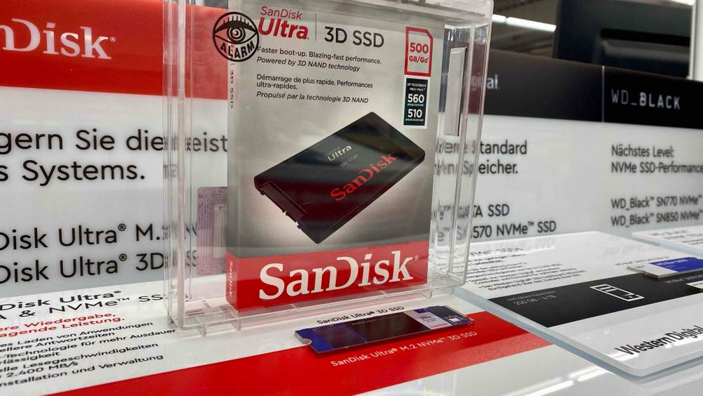 Im Test: Eine verpackte SSD (500GB) in einer transparenten Box steht hinter einer SSD die auf einer Infotafel befestigt ist.