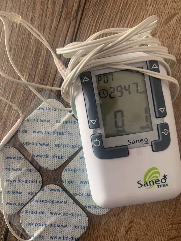 Ein weißes Saneo Tens Reizstromgerät im Test, mit Blick auf das eingeschaltete LCD, befindet sich inklusive Kabel und vier Pads auf einer Holzfläche.