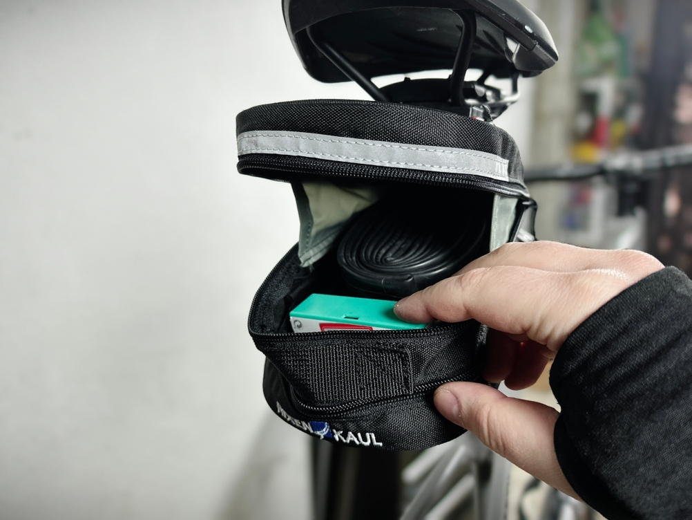 Die KLICKfix Micro 200 Satteltasche ist geöffnet und mit einem Ersatzschlauch sowie weiterem Zubehör befüllt. Eine Hand greift nach dem Inhalt.