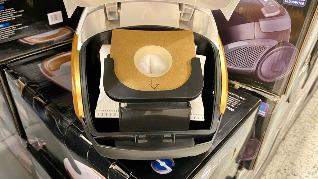 Detailansicht der Staubsaugerbeutel-Halterung in einem Bodenstaubsauger, der auf Verpackungskartons von Staubsaugern steht.