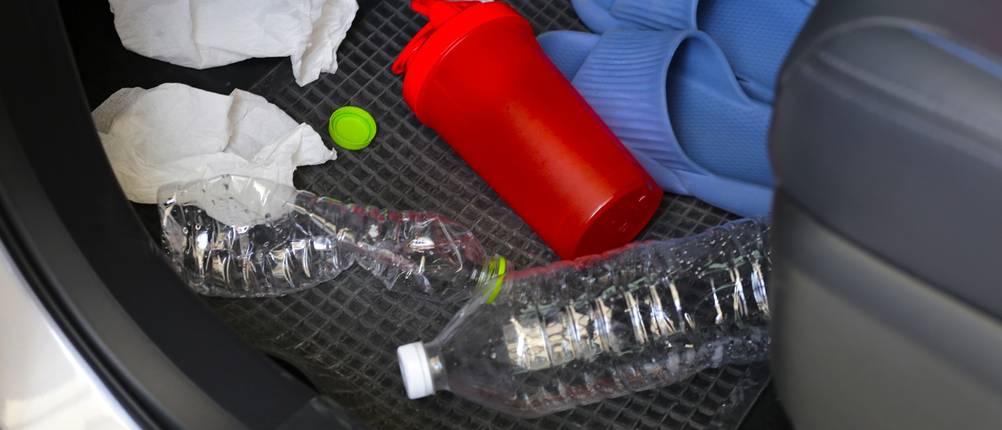 Im Fußraum eines Autos liegen leere Flaschen, eine rote Plastikflasche, blaue Schlappen und mehrere benutzte Taschentücher.