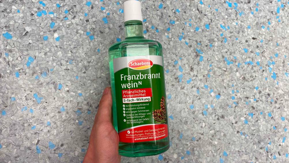 Franzbranntwein getestet: Eine Hand hält eine Flasche vor eine blau-graue Fläche.