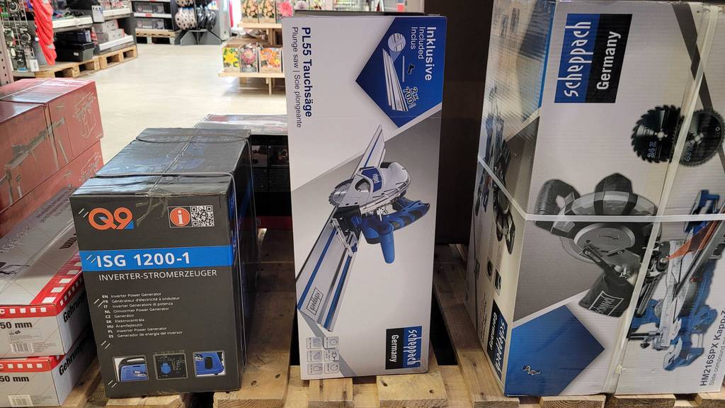 Tauchsägen mit Führungsschiene im Test: eine Originalverpackung von einem blau-schwarzen Modell der Marke Scheppach steht zwischen verpacktem Inverter-Stromerzeuger und Kappsäge auf einer Holzpalette im Baumarkt.