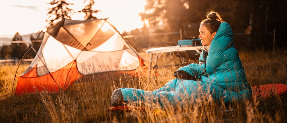 Schlafsack-Test: Frau sitzt mit türkisfarbenem Schlafsack auf einer Wiese. Im Hintergrund stehen ein Zelt und ein Campingtisch.