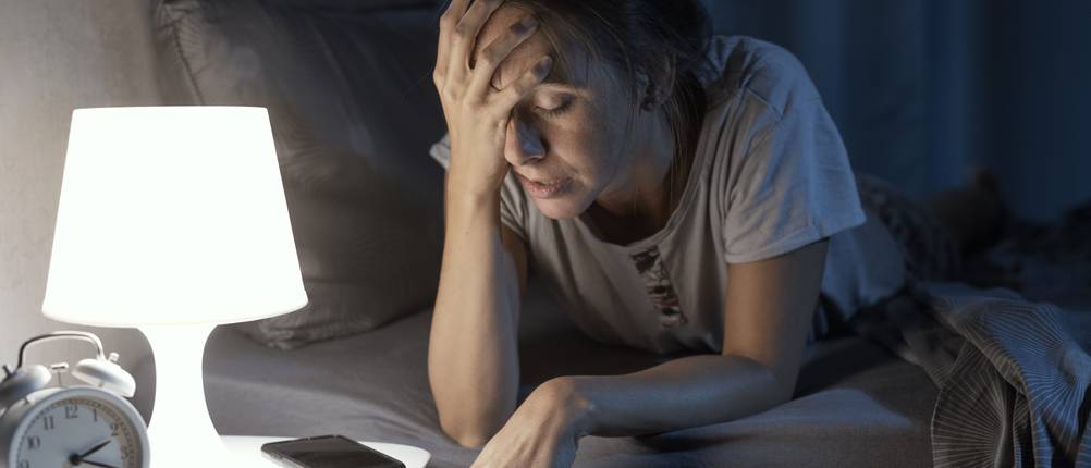 Eine Frau liegt im Bett und fasst sich verzweifelt an den Kopf.