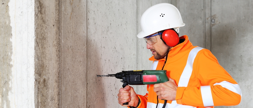 Ein Bauarbeiter in orangefarbener Warnkleidung und weißem Schutzhelm bohrt mit einer Schlagbohrmaschine in eine Betonwand