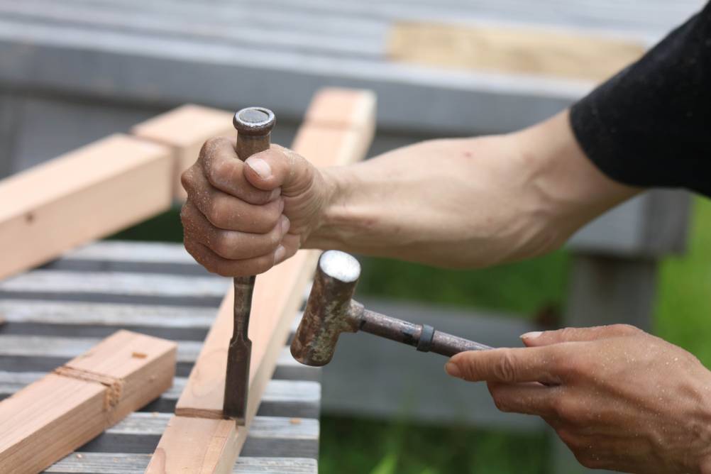 Schleifhilfe-Stechbeitel im Test: zwei Männerarme mit Stechbeitel und Hammer bearbeiten eine Aussparung in einem Kantholz.