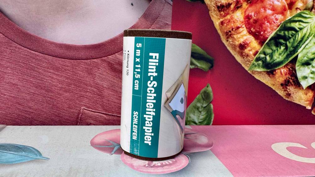 Die getestete Schleifpapier-Rolle steht vor einem Plakat mit Pizza.