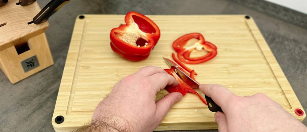 Eine rote Paprika wird auf dem Redsalt Bambusbrett mit einem kleinen Messer in Streifen geschnitten. Zwei Hälften der Paprika liegen daneben.