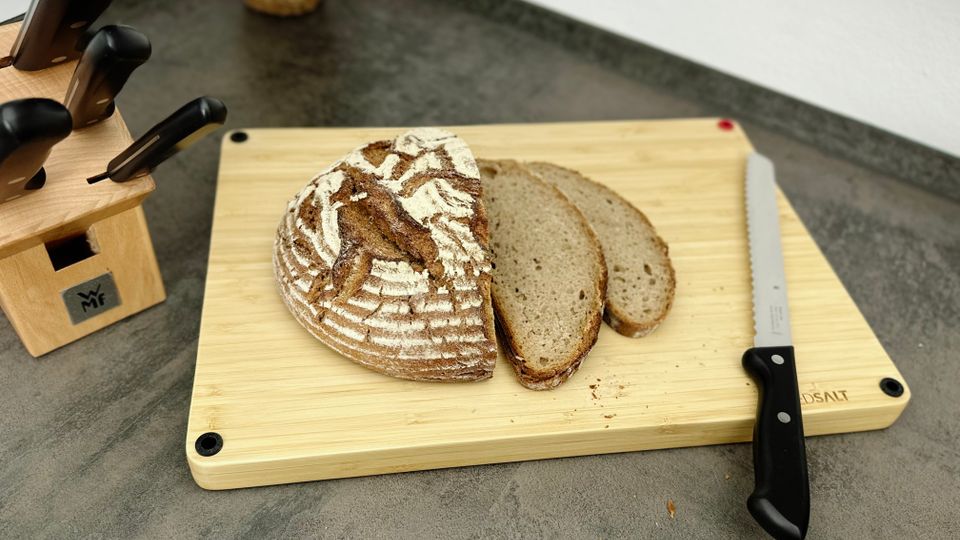 Ein rundes Bauernbrot liegt angeschnitten auf dem Redsalt Bambusbrett. Zwei Scheiben sind bereits geschnitten, daneben ein Brotmesser.