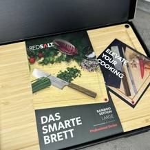 Das Redsalt Bambusbrett liegt geöffnet in der Verpackung mit Broschüre. Die Vorderseite zeigt Messer, Gemüse und das Produktlogo.