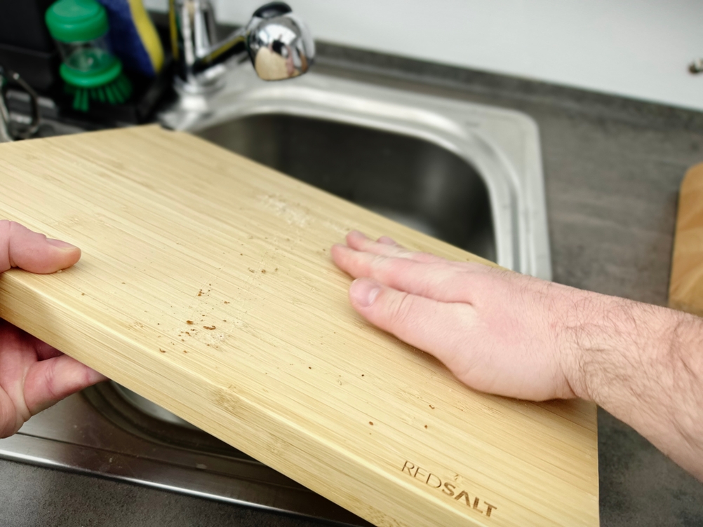 Ein Nutzer wischt mit der Hand das Redsalt-Bambusbrett über dem Spülbecken ab. Am Brett befinden sich noch Krümel.