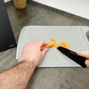 Eine Hand schneidet Karotten in Scheiben auf dem Silit-Schneidebrett 21.4223.5354. Die Oberfläche zeigt leichte Schnittspuren.