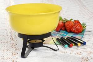 Schokofondue-Test: Ein gelbes Schokofondue mit Stövchen und Teelicht ist neben Fonduegabeln und Erdbeeren auf einem Tischtuch platziert. 