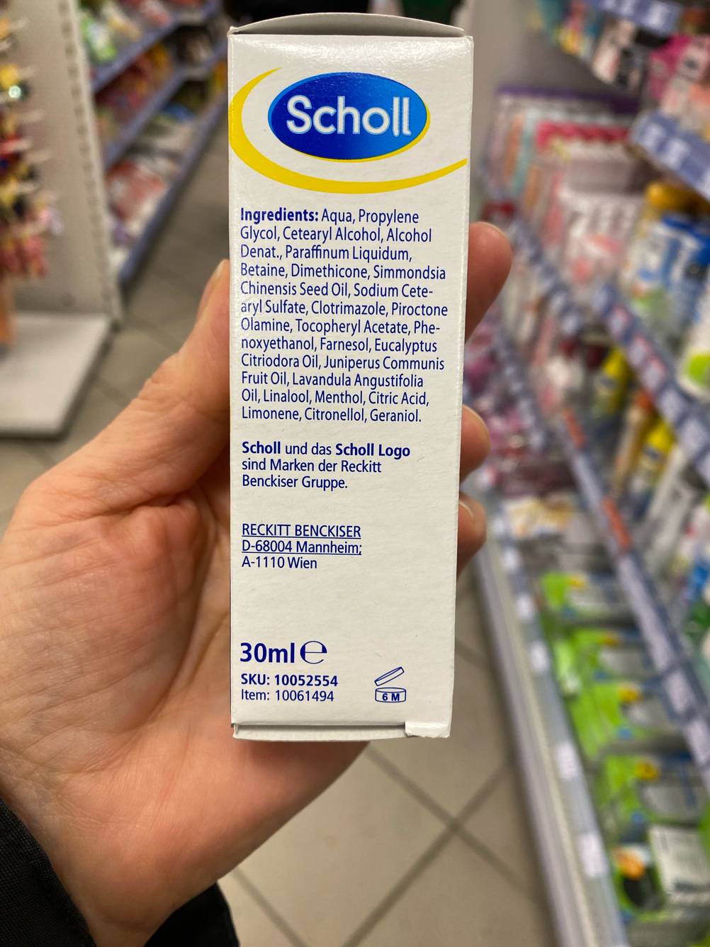 Hand hält eine Verpackung mit Scholl Fußpilz Creme im Test in einem Verkaufsbereich.