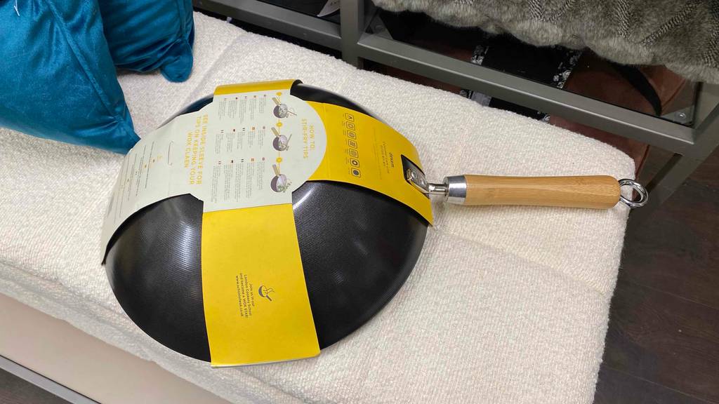 Grauer Wok im Test mit Holzgriff liegt mit der Bodenseite nach oben in der Originalverpackung auf hellem Stoff in einem Verkaufsbereich.