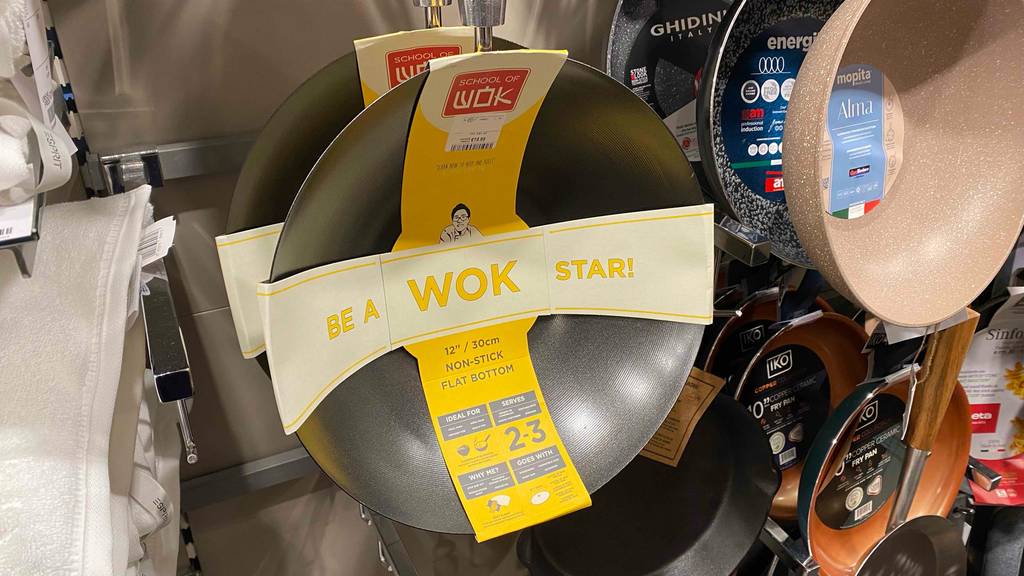 Wok im Test der Marke Wok of School hängt an einem Verkaufsstand neben anderen Pfannen und Woks.