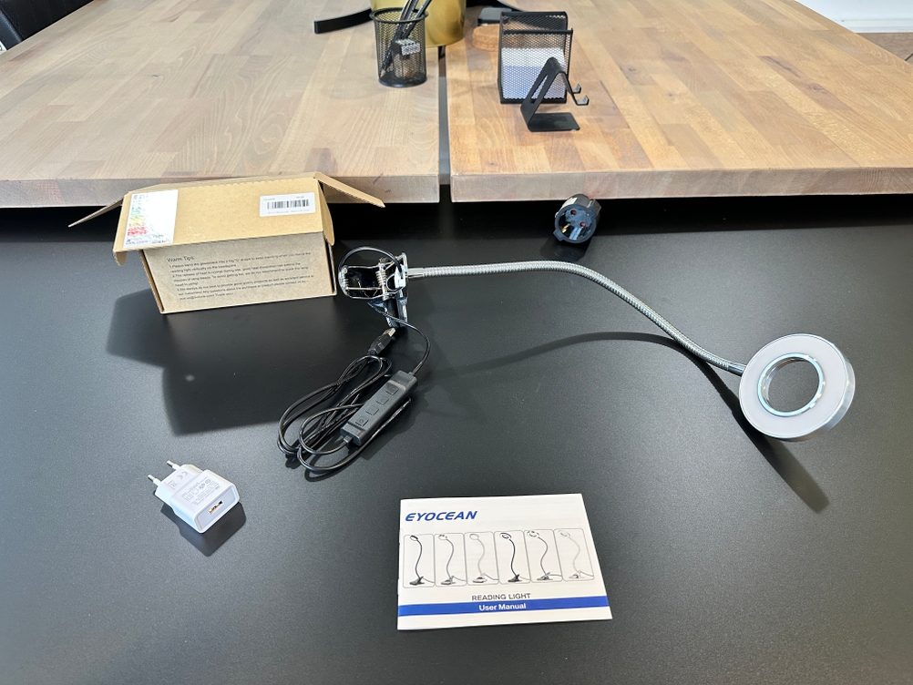 Der komplette Lieferumfang der Eyocean-LED-Leselampe, bestehend aus Lampe, USB-Netzteil, Bedienungsanleitung und Verpackung, ist auf dem Tisch ausgebreitet.