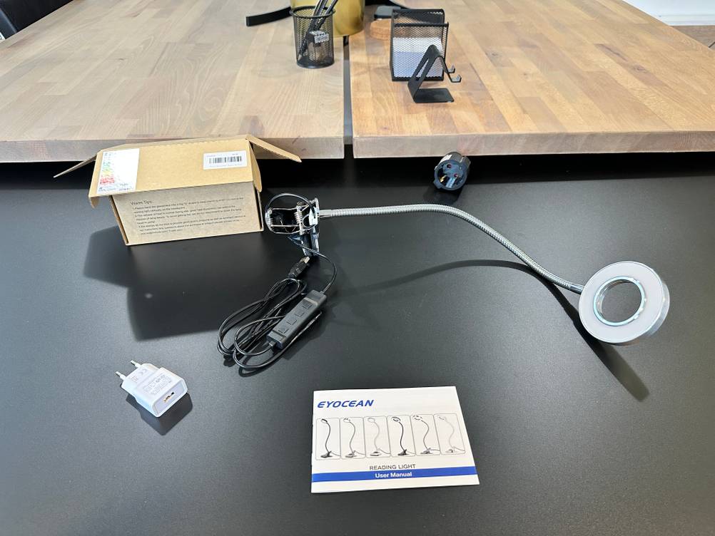 Der komplette Lieferumfang der Eyocean-LED-Leselampe, bestehend aus Lampe, USB-Netzteil, Bedienungsanleitung und Verpackung, ist auf dem Tisch ausgebreitet.