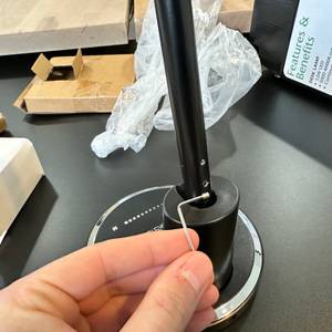 Eine Hand setzt den Innensechskantschlüssel an der Verschraubung zwischen Lampenfuß und Stange der Jukstg HD1906C an.