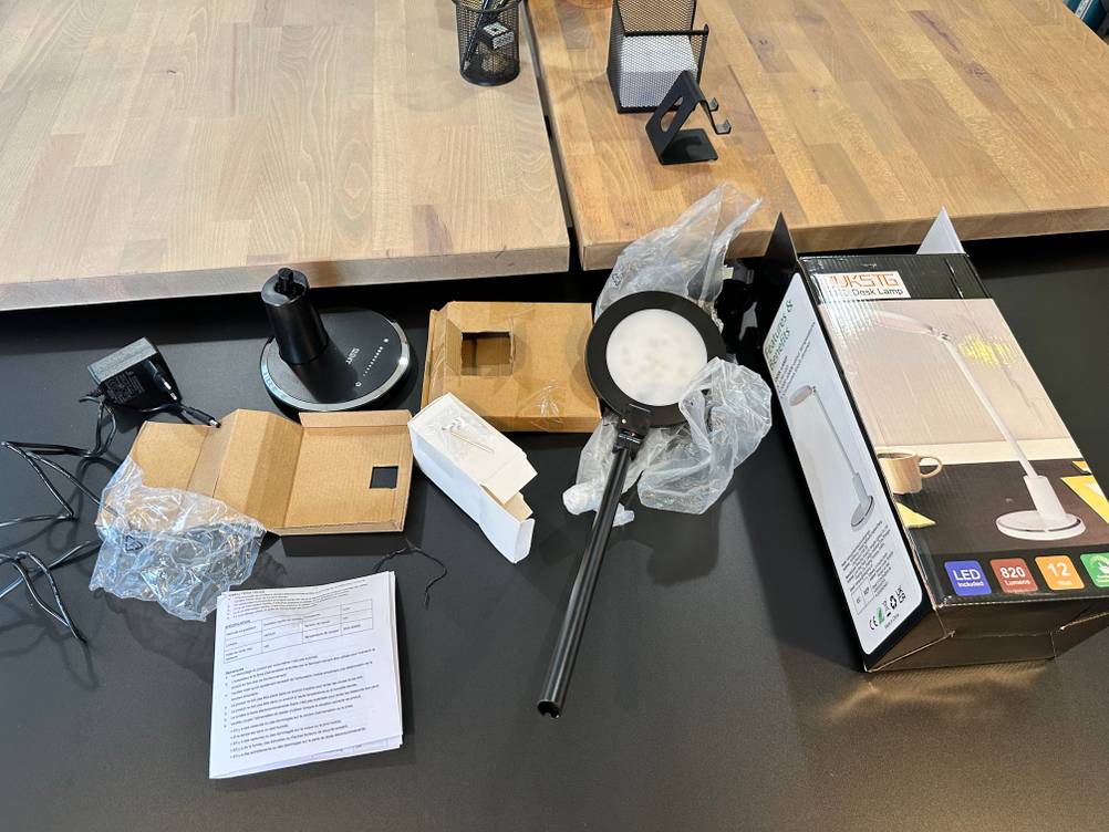 Der komplette Lieferumfang der Jukstg-HD1906C-Schreibtischlampe, bestehend aus Lampenkopf, Lampenfuß, Netzadapter, Montagematerial, Anleitung und Verpackung, liegt ausgelegt auf dem Tisch.