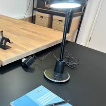 Die vollständig aufgebaute und eingeschaltete Jukstg-HD1906C-Schreibtischlampe beleuchtet einen Collegeblock auf einem schwarzen Schreibtisch.