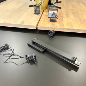 Die eingeklappte Lepro-LED-Schreibtischlampe und das zugehörige Netzkabel liegen auf einem schwarzen Tisch vor einer Holzarbeitsfläche.
