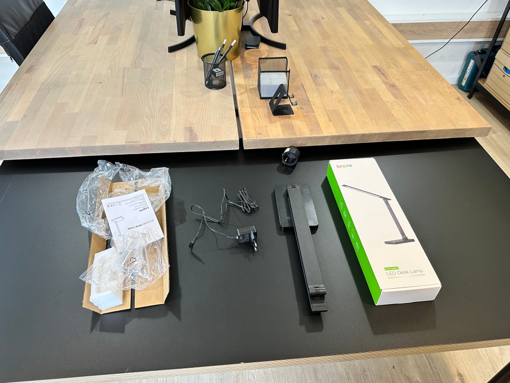 Der vollständige Lieferumfang der Lepro-LED-Schreibtischlampe, bestehend aus Lampe, Netzadapter, Anleitung und Verpackungsmaterial, ist auf dem Tisch ausgelegt.