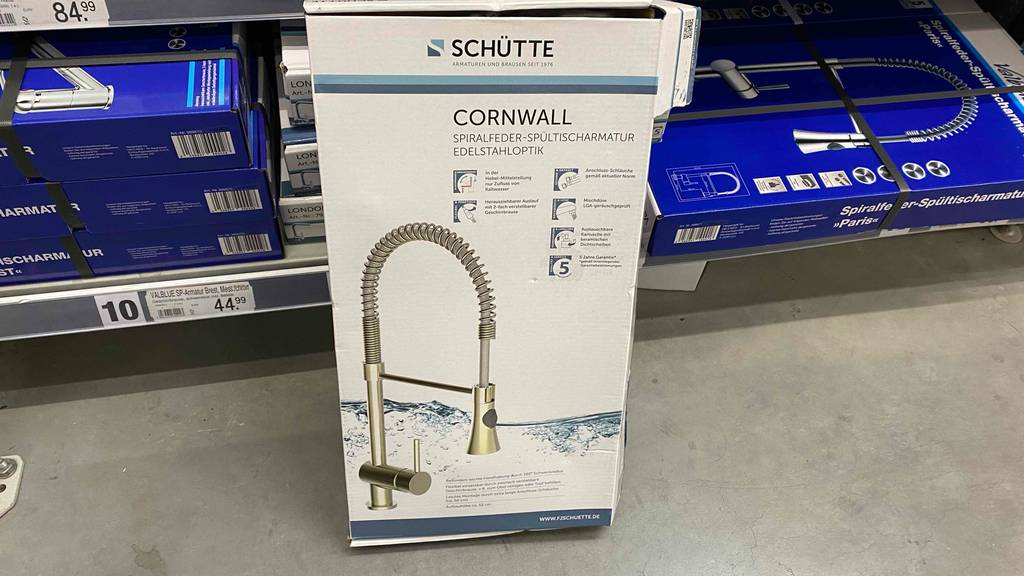 Eine Original-Verpackung inklusive getesteter Küchenarmatur in Edelstahloptik der Marke Schütte steht an einen Verkaufsbereich gelehnt auf einem grauen Steinboden.