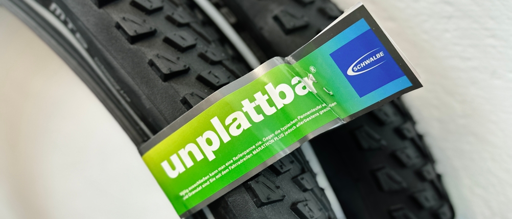 Verpackungslabel mit der Aufschrift „unplattbar“ am Schwalbe Marathon Plus MTB HS468. Der Reifen liegt zusammengefaltet im Hintergrund.