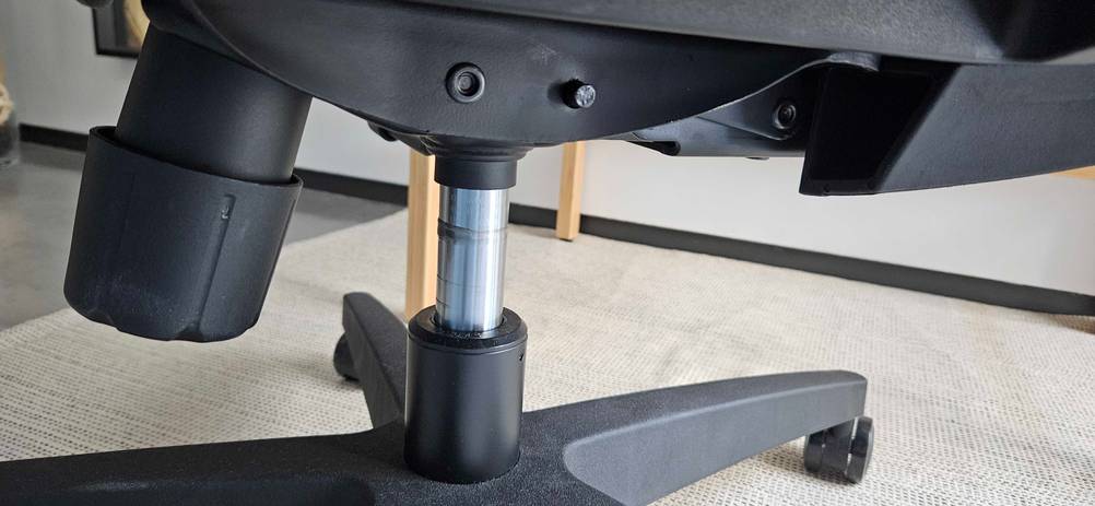 Nahaufnahme der Hydraulikmechanik zum Verstellen der Sitzhöhe eines schwarzen getesteten Bürostuhls auf hellem Teppichboden.