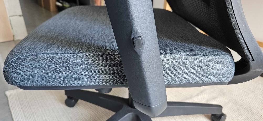 Detailansicht eines schwarz-weiß meliert bezogenen Bürostuhls im Test, mit Blick auf den Knopf zum Höhen-Verstellen der Armlehne.