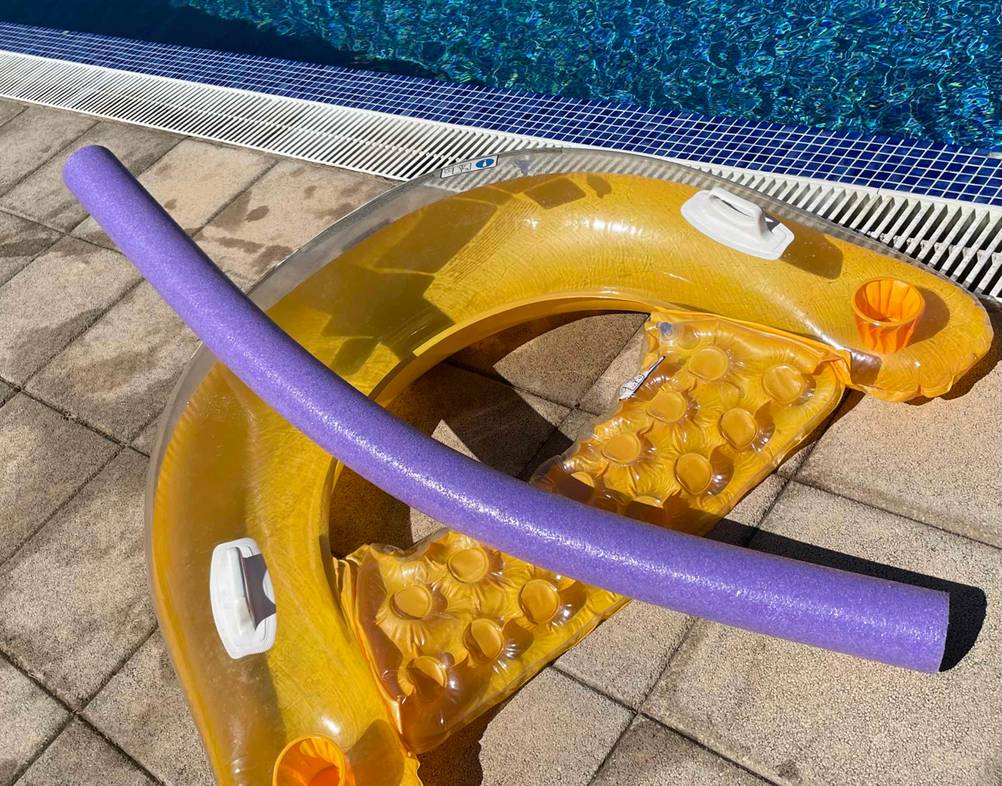 Lilafarbige getestete Schwimmnudel liegt auf einer gelben Schwimmmatratze am Pool-Beckenrand.