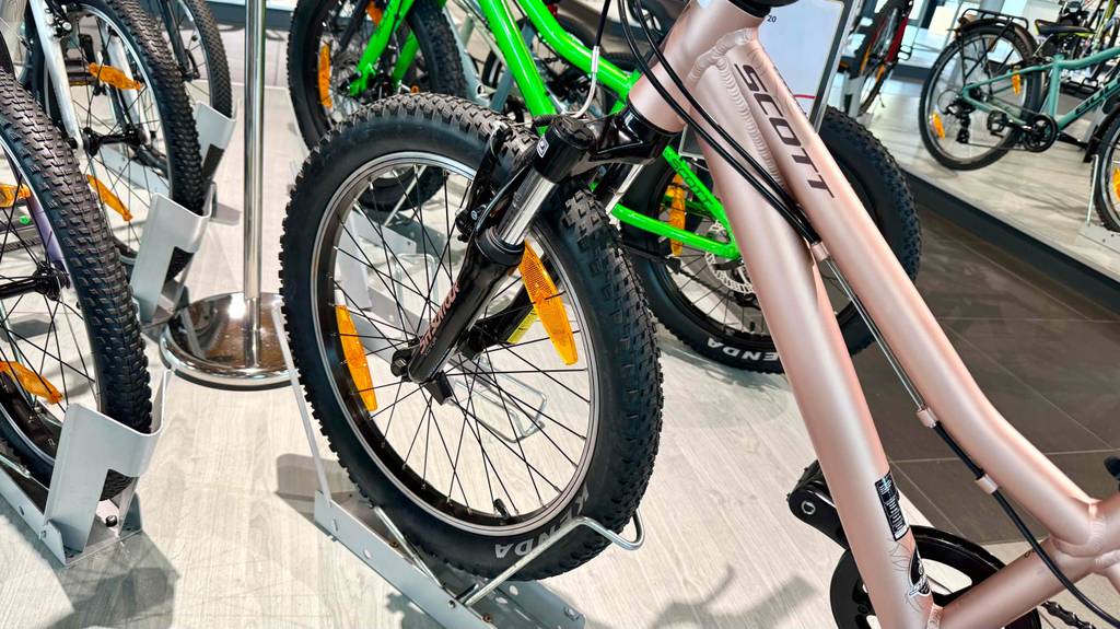 Das rosafarbene getestete Mountainbike 20 Zoll für Damen steht mit Fokus auf den Vorderreifen auf einem Ausstellungspodest neben weiteren Fahrrädern.