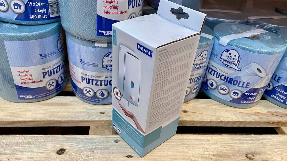 Ein getesteter Wand-Seifenspender befindet sich verpackt vor mehreren Rollen Putztüchern.