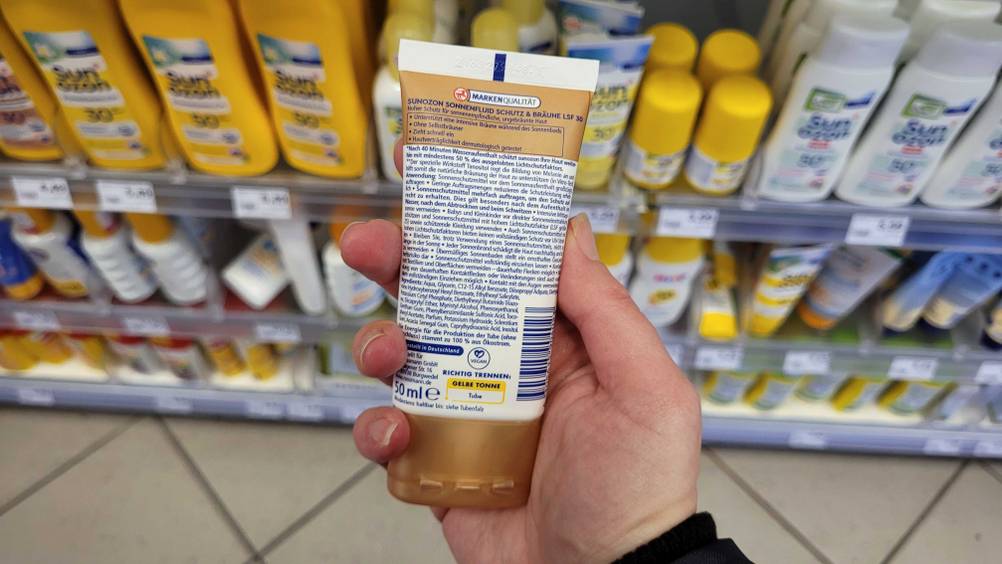 Die Rückseite einer Tube getesteter Selbstbräuner fürs Gesicht von einer Hand vor ein Regal gehalten.