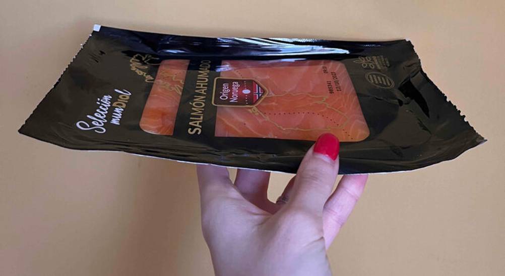 Eine Damenhand hält eine Verpackung geschnittenen Räucherlachs im Test vor einer beigefarbenen Wand.