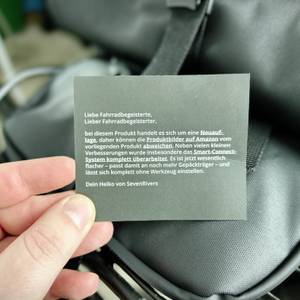 Ein Hinweiszettel mit Infos zur Sevenrivers-Fahrradtasche wird vor der montierten Tasche in der Hand gehalten. Der Text erklärt eine Produktüberarbeitung.
