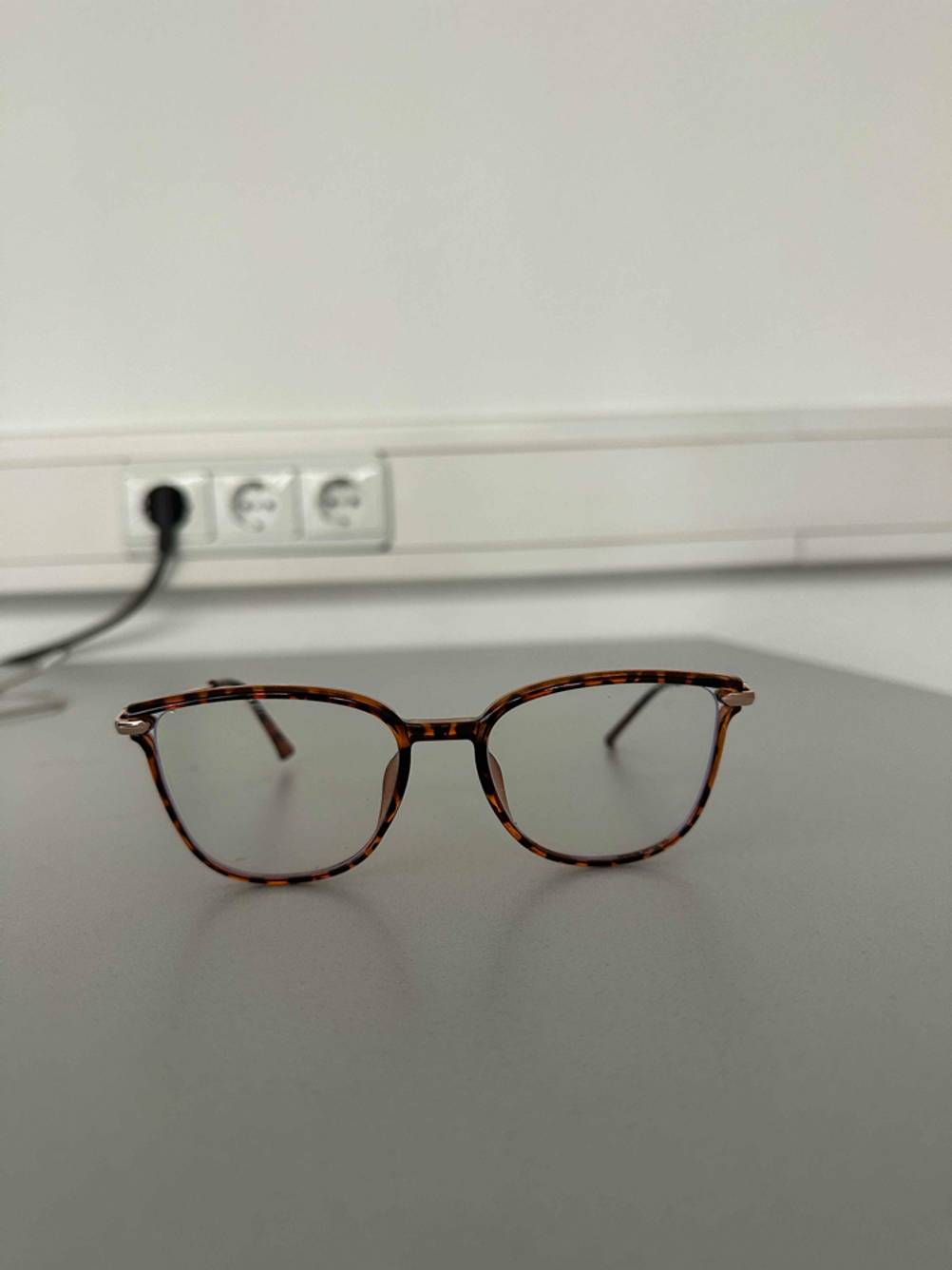 Eine Blaulichtfilter-Brille im Test mit braun gemustertem Vollrahmen liegt auf einer grauen Oberfläche; im Hintergrund eine Steckerleiste.