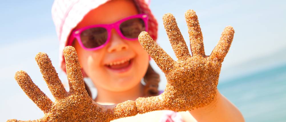Badeschuhe für Kinder im Test: Ein Mädchen mit Sonnenbrille und Sonnenhut hält ihre sandigen Hände in die Kamera.