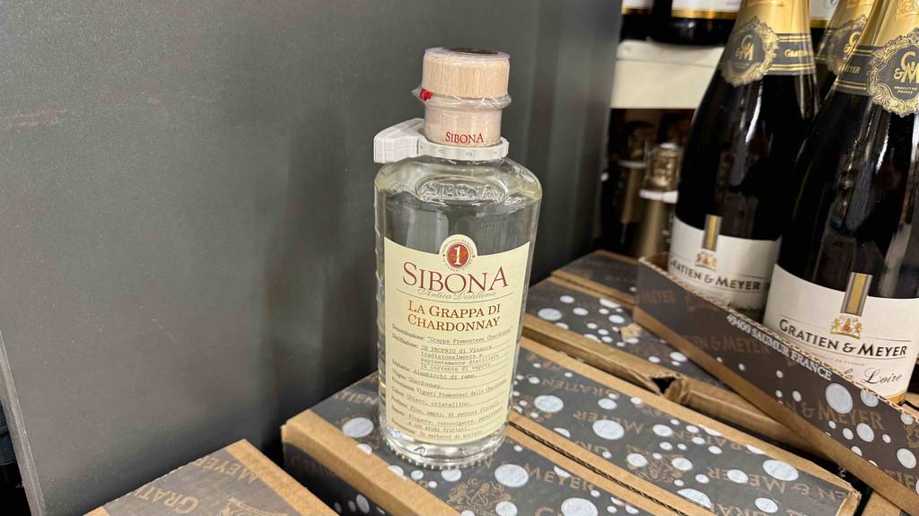 Test: Chardonnay Grappa in der Flasche inklusiven Holzkorken steht auf einem Karton neben anderen Spirituosen in der Flasche.