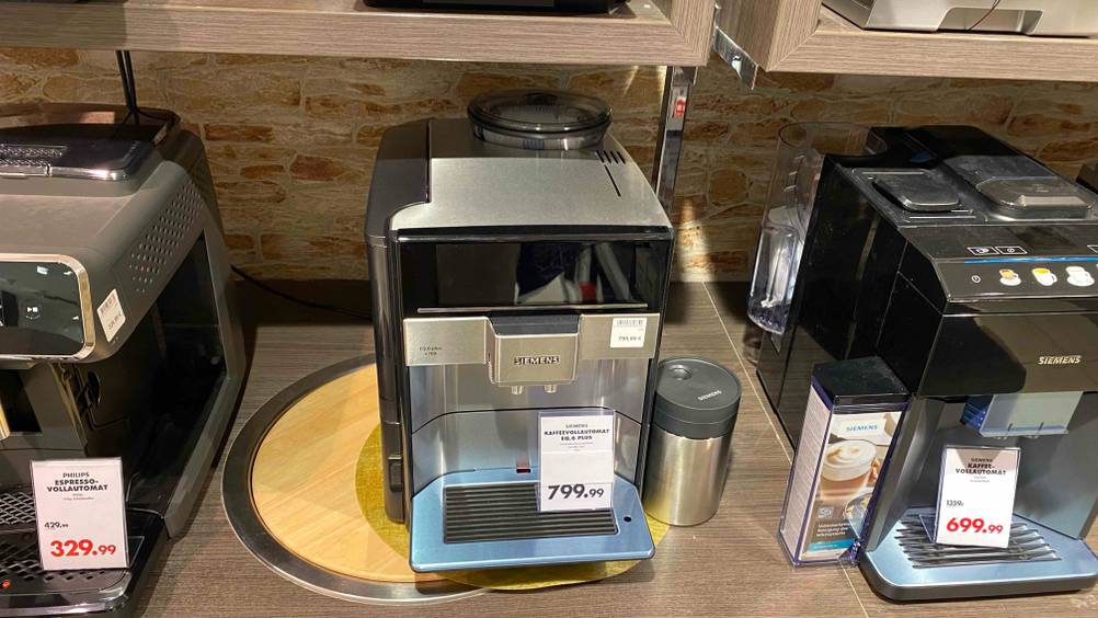 Ein getesteter Kaffeevollautomat von schräg oben in einer Verkaufsausstellung neben anderen Geräten und Preisangaben.