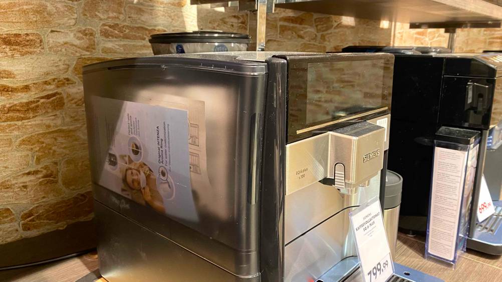 Ein getesteter Kaffeevollautomat von der Seite in einer Ausstellungsfläche mit einem Preisschild davor, im Hintergrund andere Produkte.