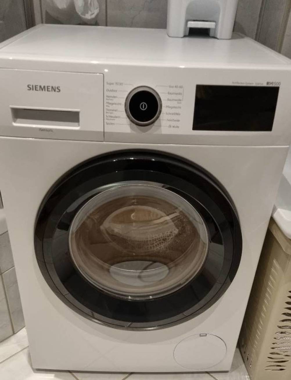 Eine weiße getestete Siemens-Waschmaschine mit dunkler Tür steht ausgeschaltet in einem Badezimmer.