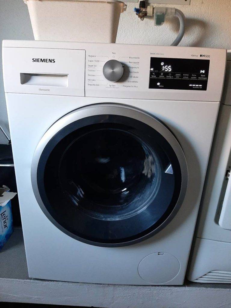 Eine weiße getestete Siemens-Waschmaschine steht mit eingeschaltetem Betrieb und Display in einem Waschraum.