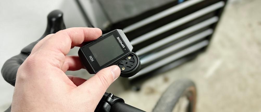 Eine Hand hält das Sigma Sport ROX 4.0 über einer Lenkerhalterung. Das Display zeigt keine aktiven Daten an.