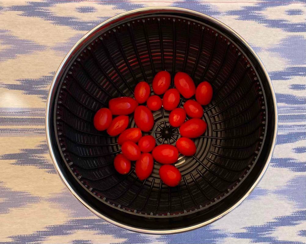 Ansicht von oben auf eine Salatschleuder aus Edelstahl im Test, die inklusive schwarzem Korbeinsatz mit Datteltomaten auf einer lila-weißen gemusterten Tischdecke steht.